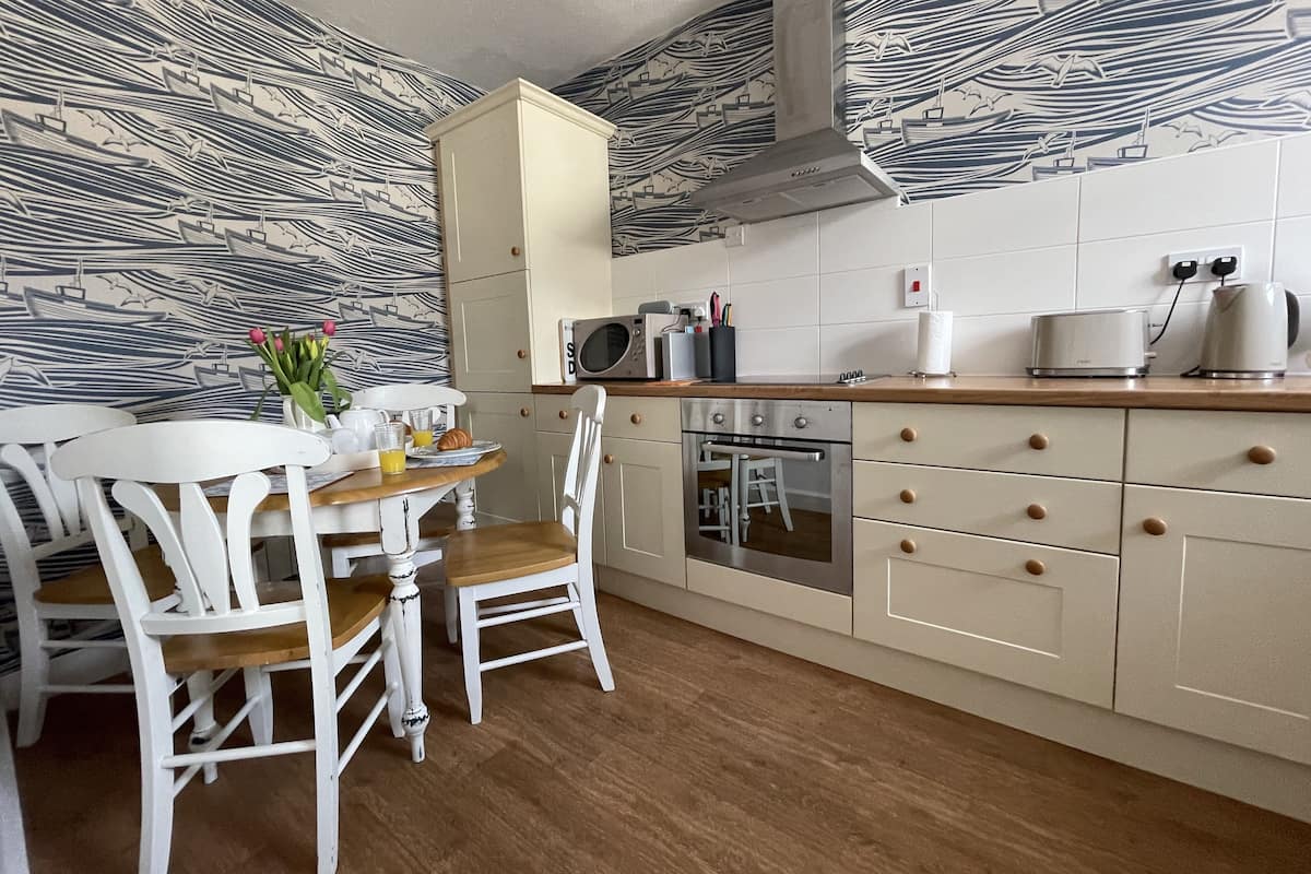 Une cuisine avec une table en bois, des chaises blanches, un four à micro-ondes, un four traditionnel et un crédence décorée.