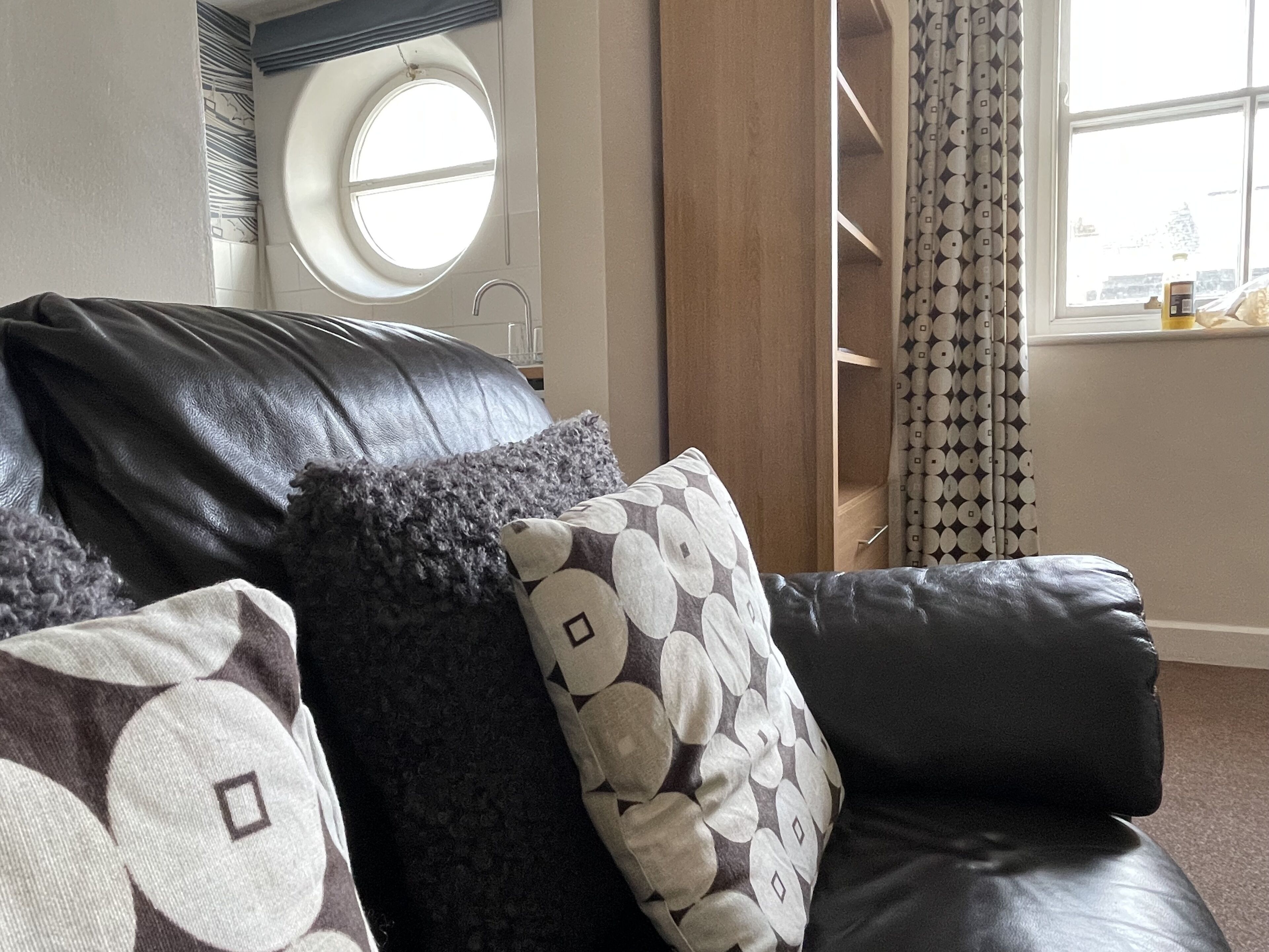 A black leather sofa with patterned cushions in a room with a window, a circular skylight, and a patterned curtain.