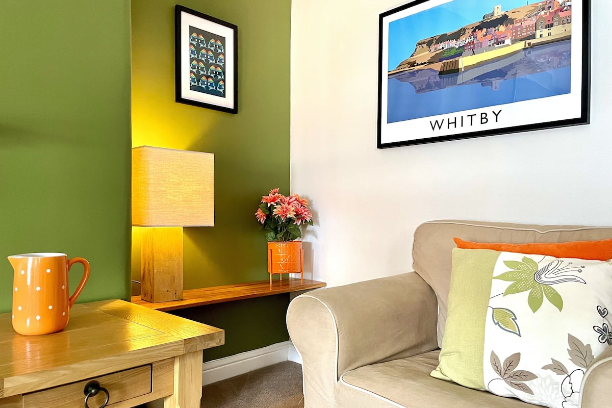 Un salon chaleureux avec une table en bois, une lampe, un vase rempli de fleurs, un tableau encadré représentant Whitby et un canapé beige agrémenté d’un coussin à motifs floraux.