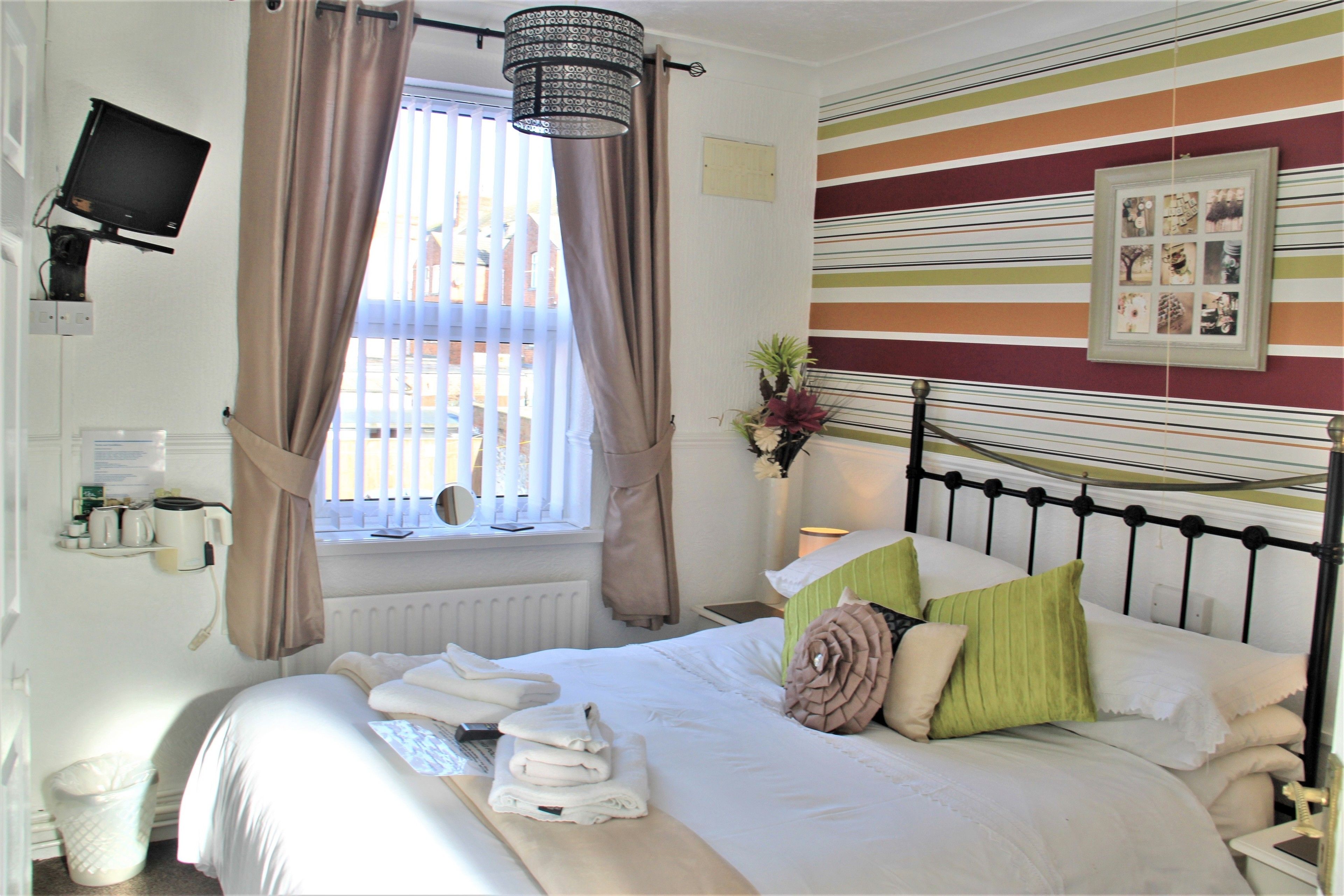 A neatly made bed with pillows, a bedside table with a lamp, a window with curtains, and a wall with a striped pattern.