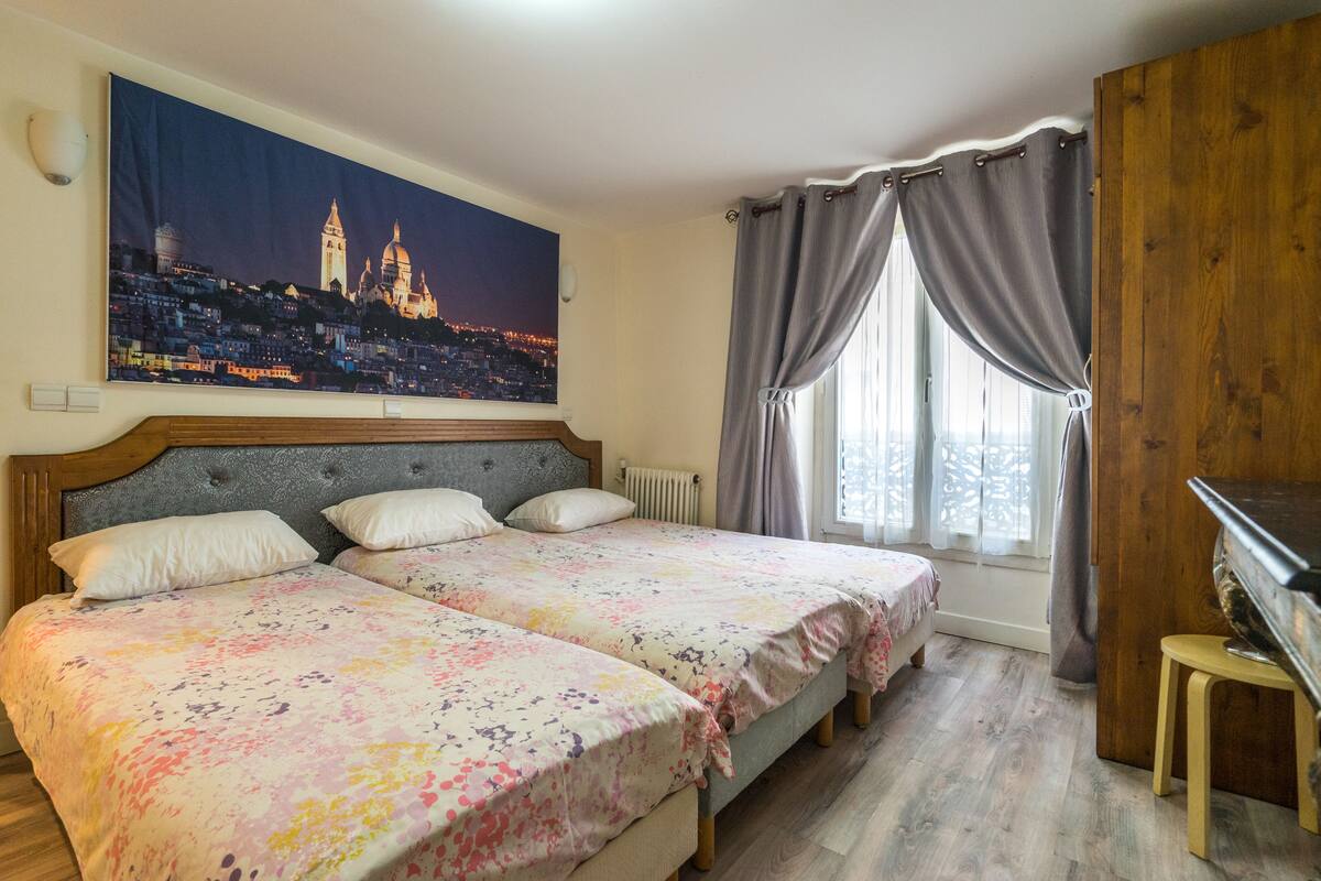 Un dormitorio con dos camas, un cuadro de gran tamaño con un paisaje urbano, un cabecero de madera, un radiador, un piano y una ventana con cortinas.