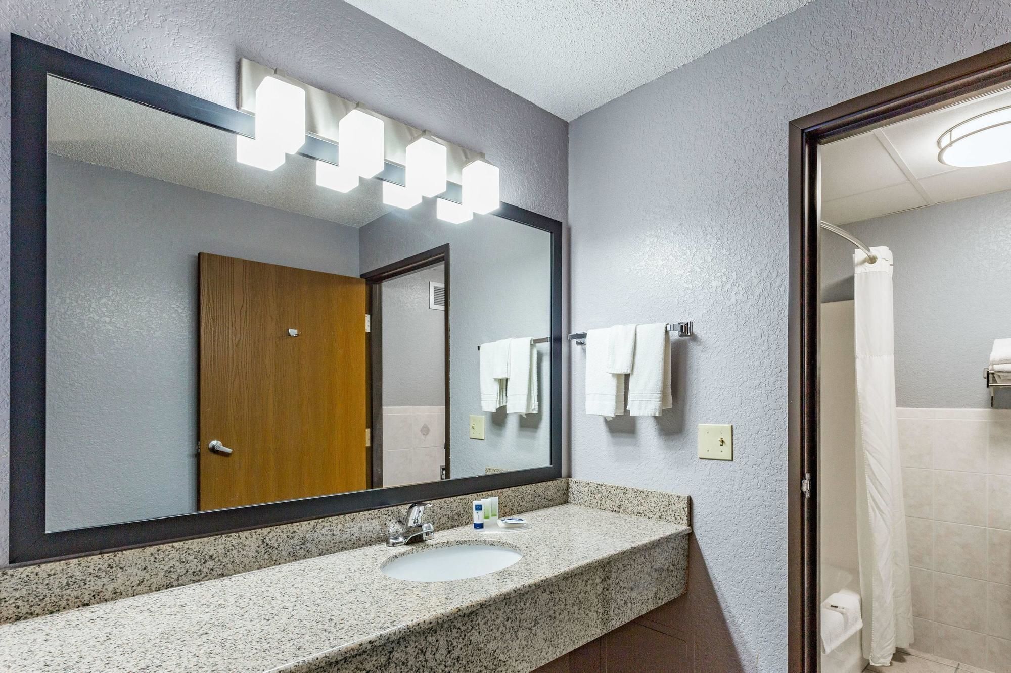 A bathroom with a granite countertop sink, a large mirror with lights, a wooden door, and a shower area.
