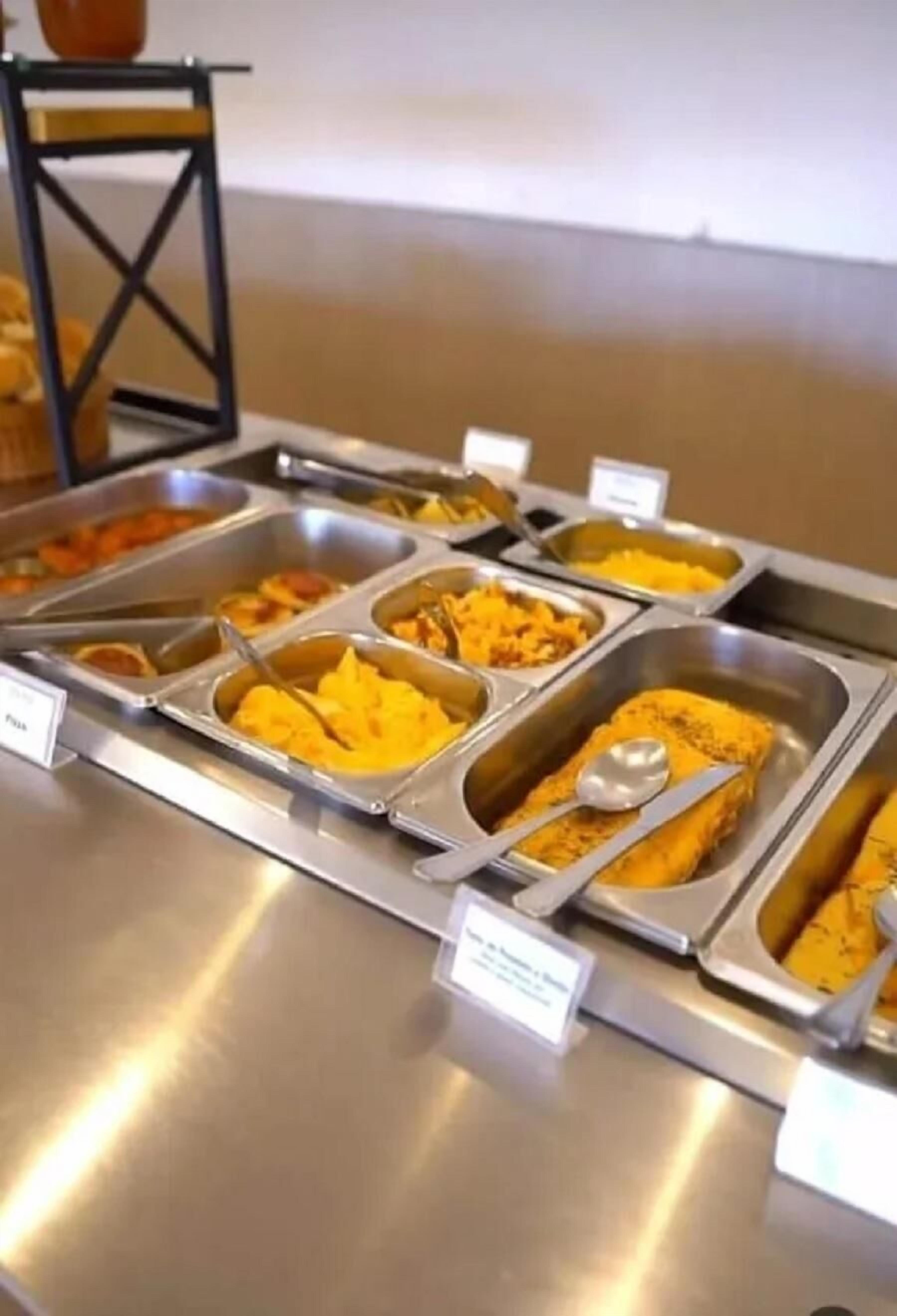 Buffet com diversos alimentos em recipientes metálicos, cada um com uma colher e placas indicativas.