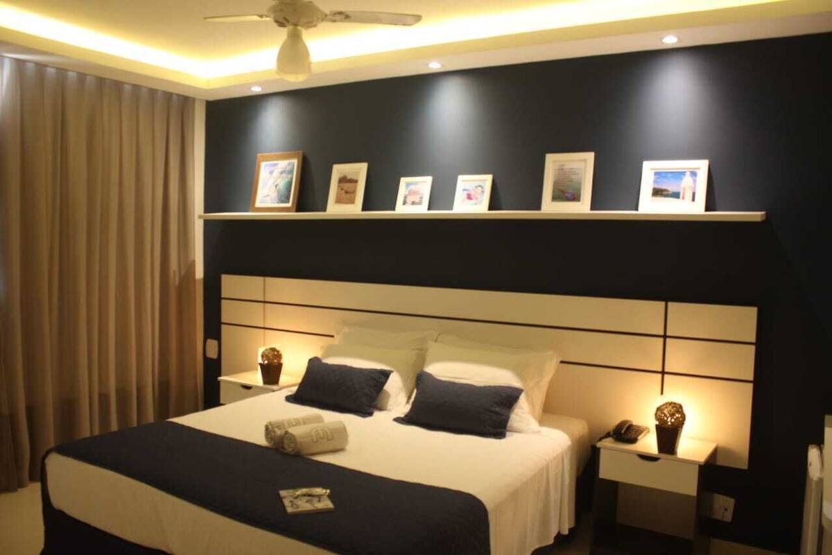 Habitación de hotel con una cama, mesitas de noche, un ventilador de techo y una repisa en la pared con fotografías enmarcadas.