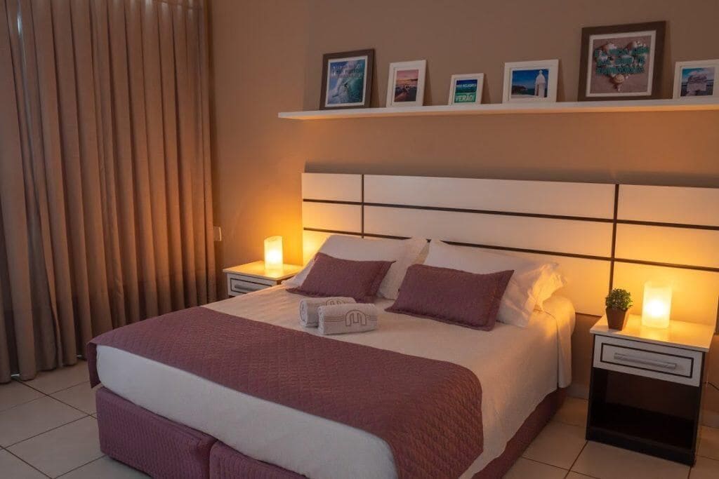 Una cama bien hecha con un cabecero, dos mesitas de noche con lámparas, y una repisa sobre la cama con cuadros enmarcados.
