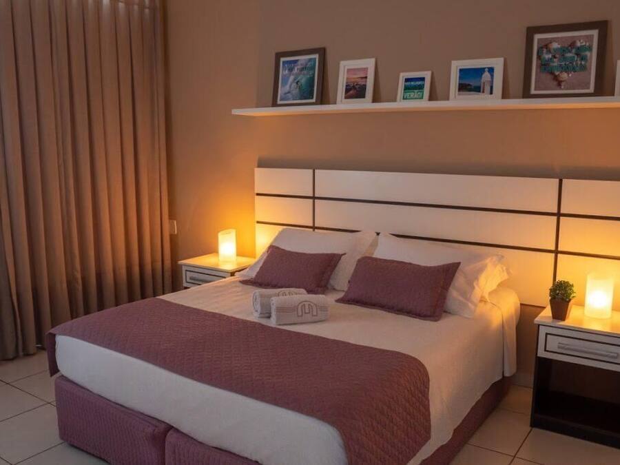 Una cama bien hecha con un cabecero, dos mesitas de noche con lámparas, y una repisa sobre la cama con cuadros enmarcados.