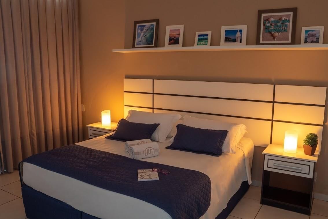 Cama bem arrumada com cabeceira, duas mesas de cabeceira com abajures e uma prateleira acima da cama exibindo fotografias emolduradas.