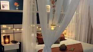 Et soveværelse med himmelseng, natbord med lampe og en vægmonteret hylde med pynteting.
