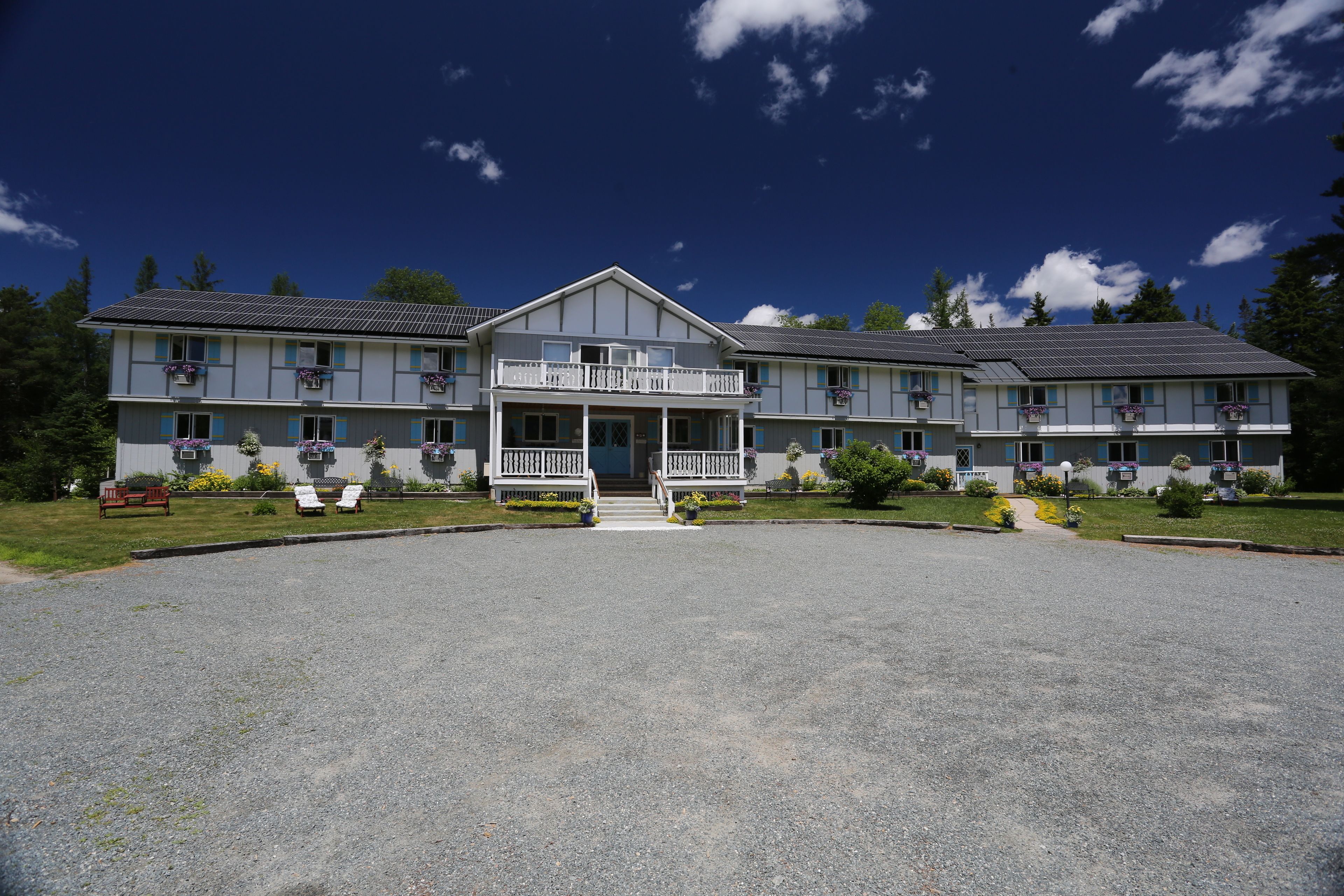 A two-story hotel with a central entrance, balconies with flower boxes, and a gravel driveway.