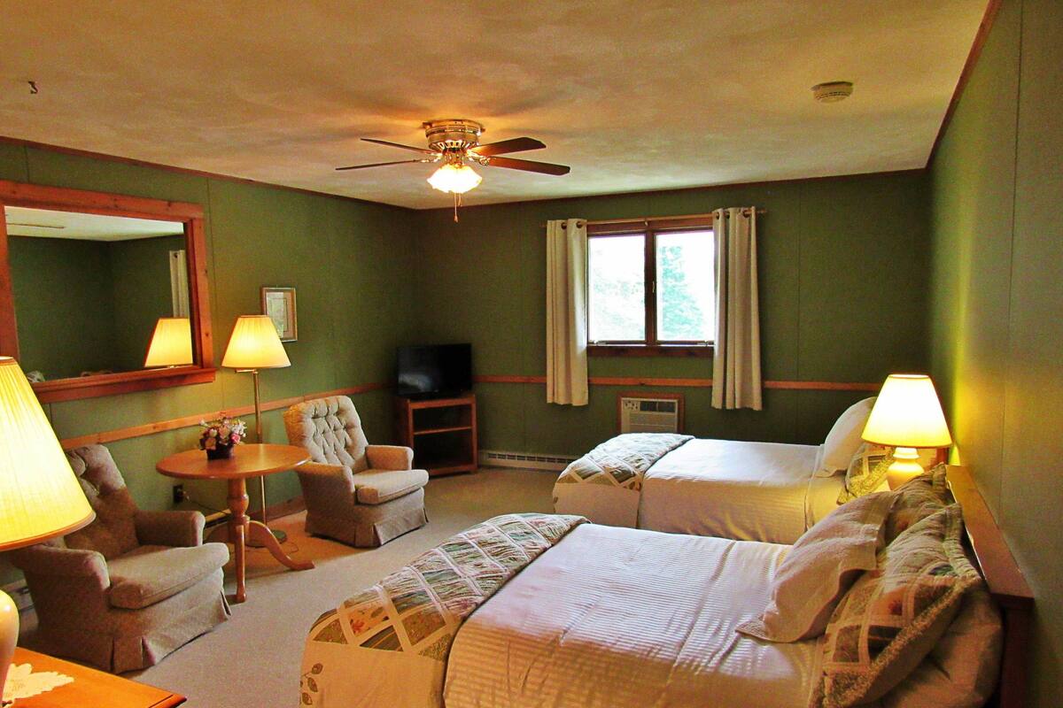 Quarto de hotel com cama, duas cadeiras, uma mesinha, ventilador de teto, televisão e janela com cortinas.