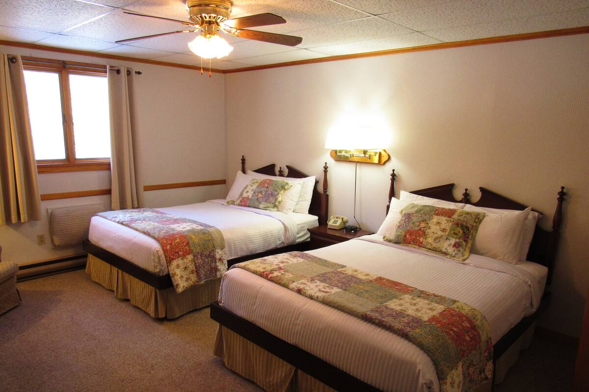 Quarto de hotel com duas camas de solteiro, um ventilador de teto, mesa de cabeceira com telefone e uma janela com cortinas.