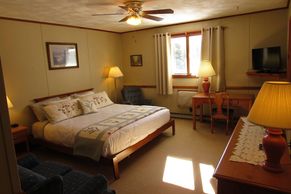 Quarto de hotel com uma cama, duas cadeiras, uma escrivaninha com abajur, televisão e ventoinha de teto.