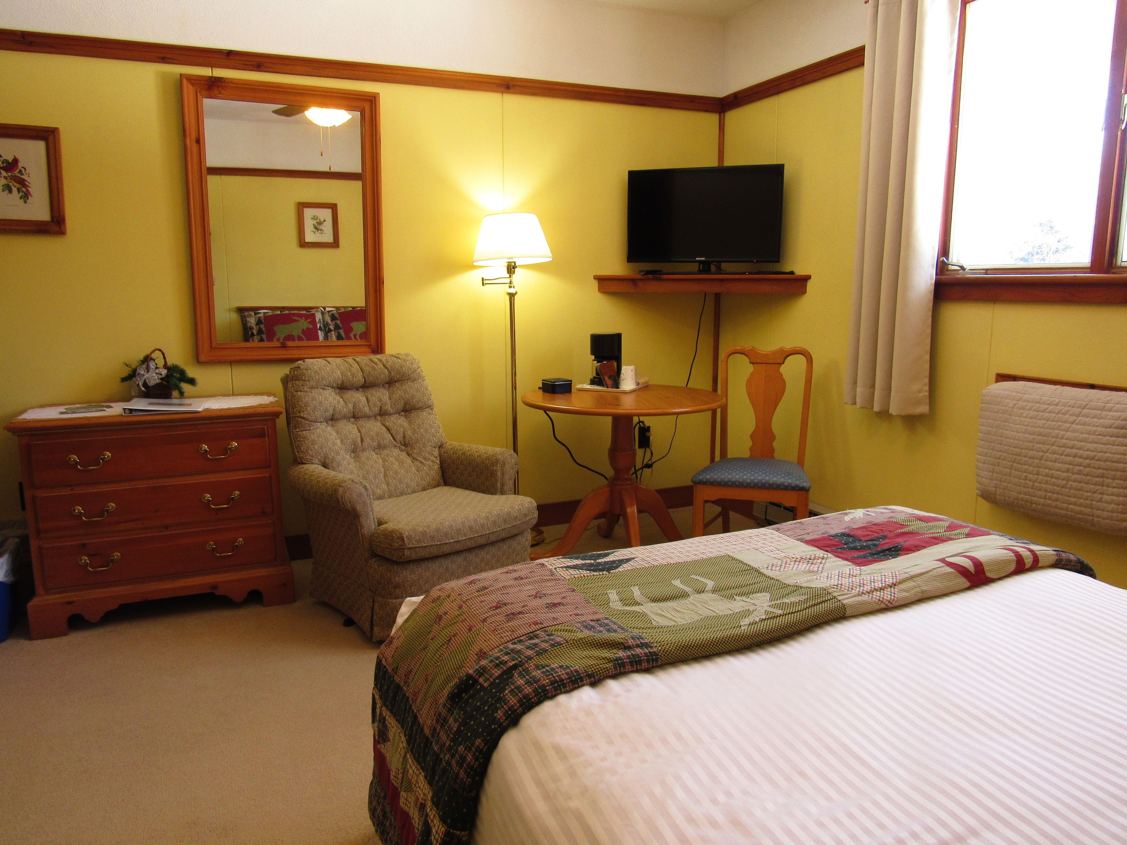 A hotel room with a bed, a wooden dresser, a chair, a desk with a lamp, a television, and a window with curtains.