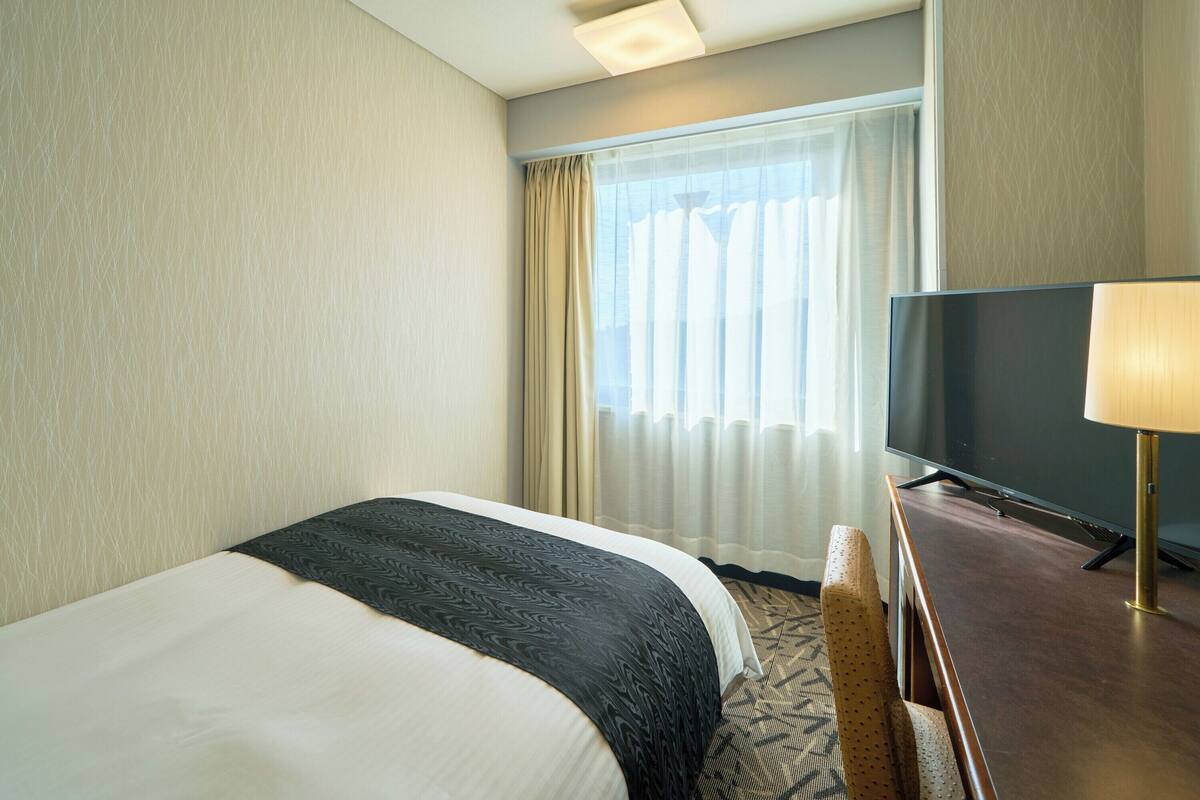 A hotel room with a bed, a television, a lamp, and a window with curtains.