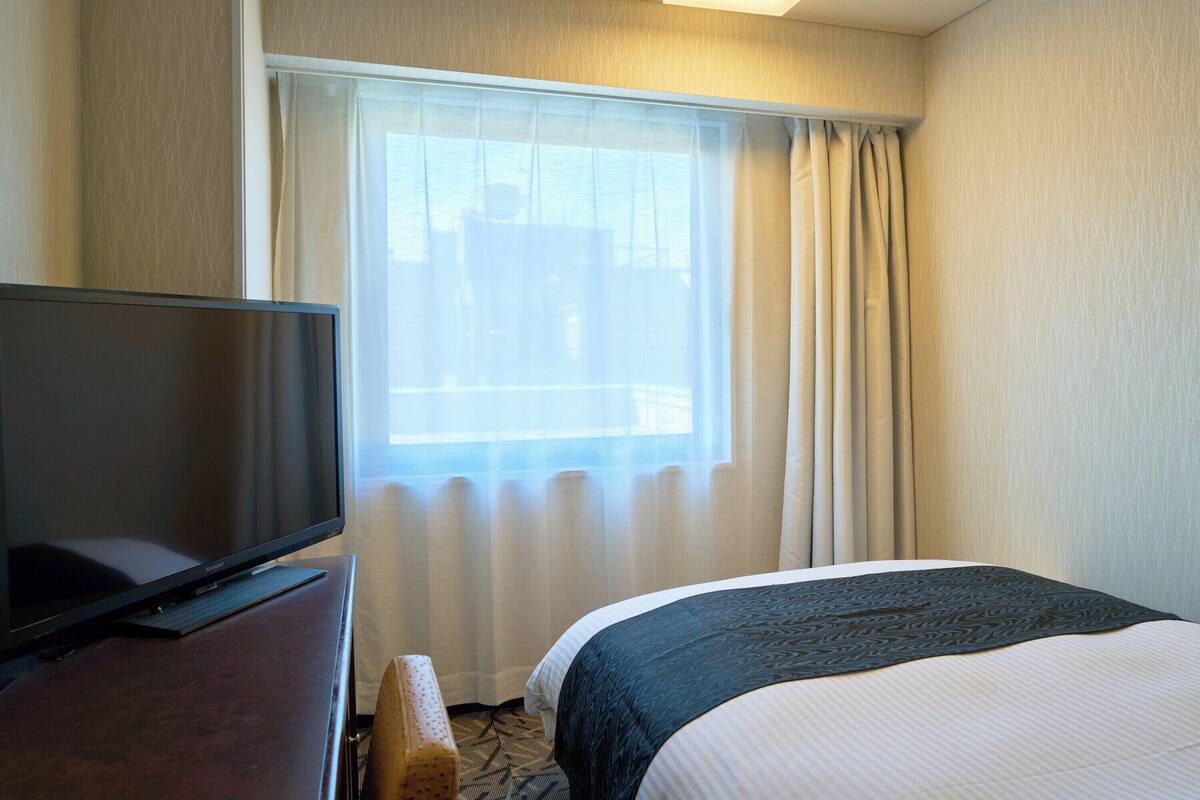 A hotel room with a bed, a television on a stand, and a window with curtains.