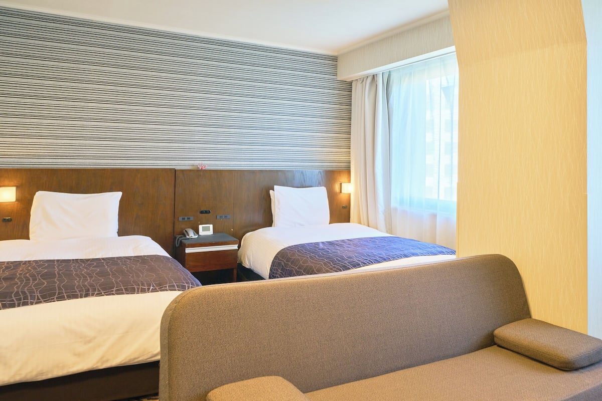 Habitación de hotel con dos camas, un sofá, una mesita de noche con teléfono y una ventana con cortinas.