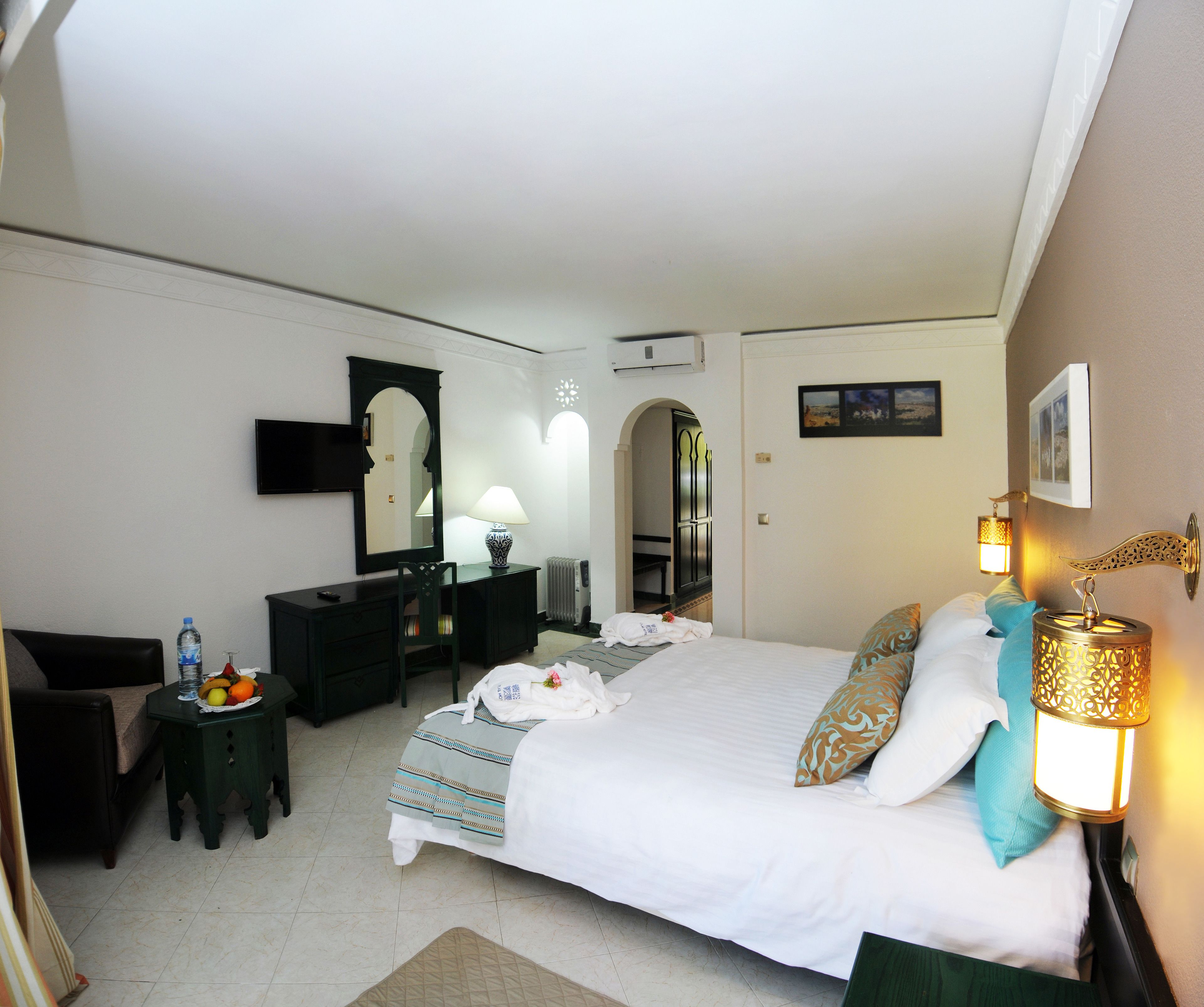 Quarto de hotel com cama, escrivaninha, cadeira, televisão, luminária e quadro na parede.