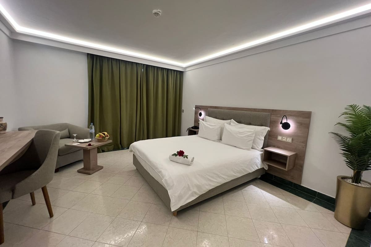 Quarto de hotel moderno com uma cama bem arrumada, uma escrivaninha com cadeira, uma mesa de cabeceira com abajur e um vaso de planta.