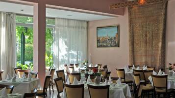 Un comedor con mesas dispuestas para el servicio, ventanas grandes con cortinas y un cuadro en la pared.