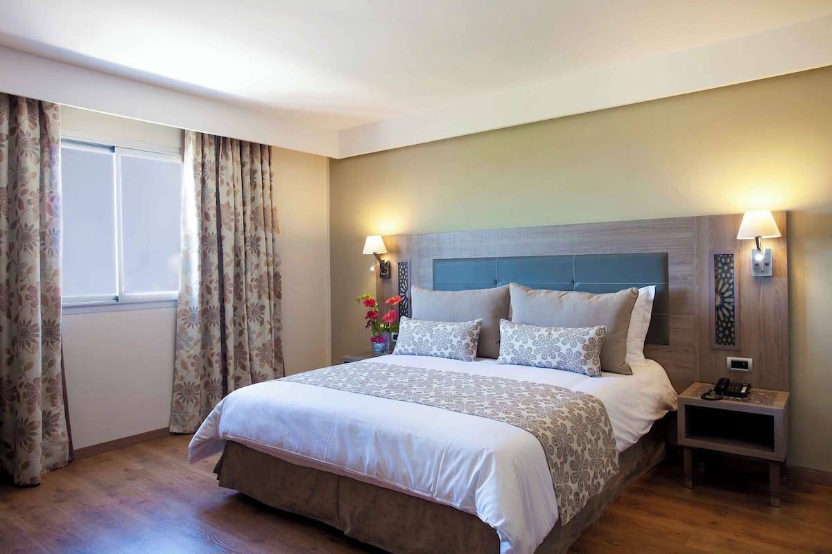 Uma cama bem arrumada com um cobre-leito estampado, uma mesa de cabeceira com abajur e uma janela com cortinas.