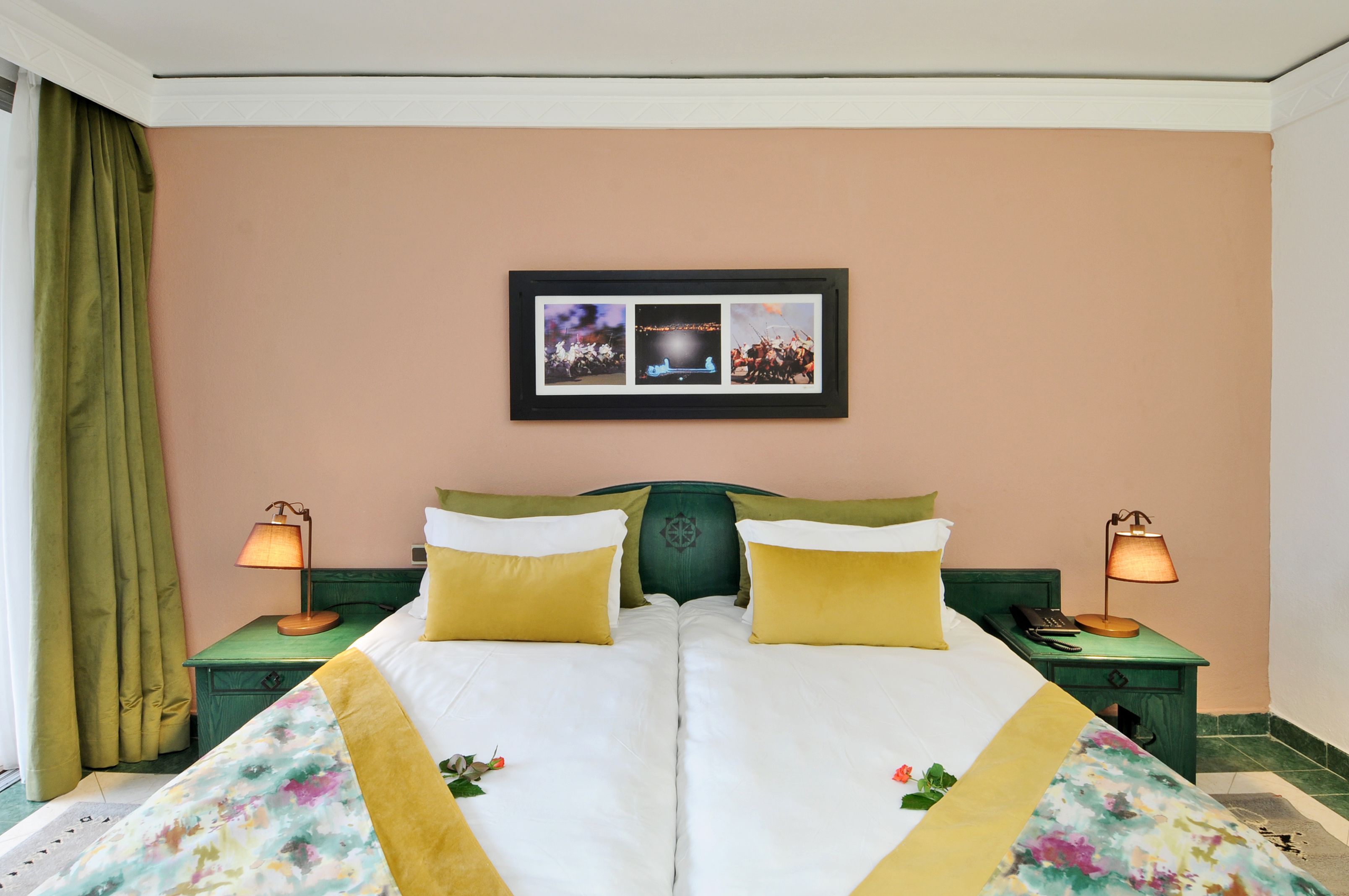 Een tweepersoonsbed met bloemenbeddengoed, twee nachtkastjes met lampen, een lijst met een afbeelding aan de muur en een groen gordijn.