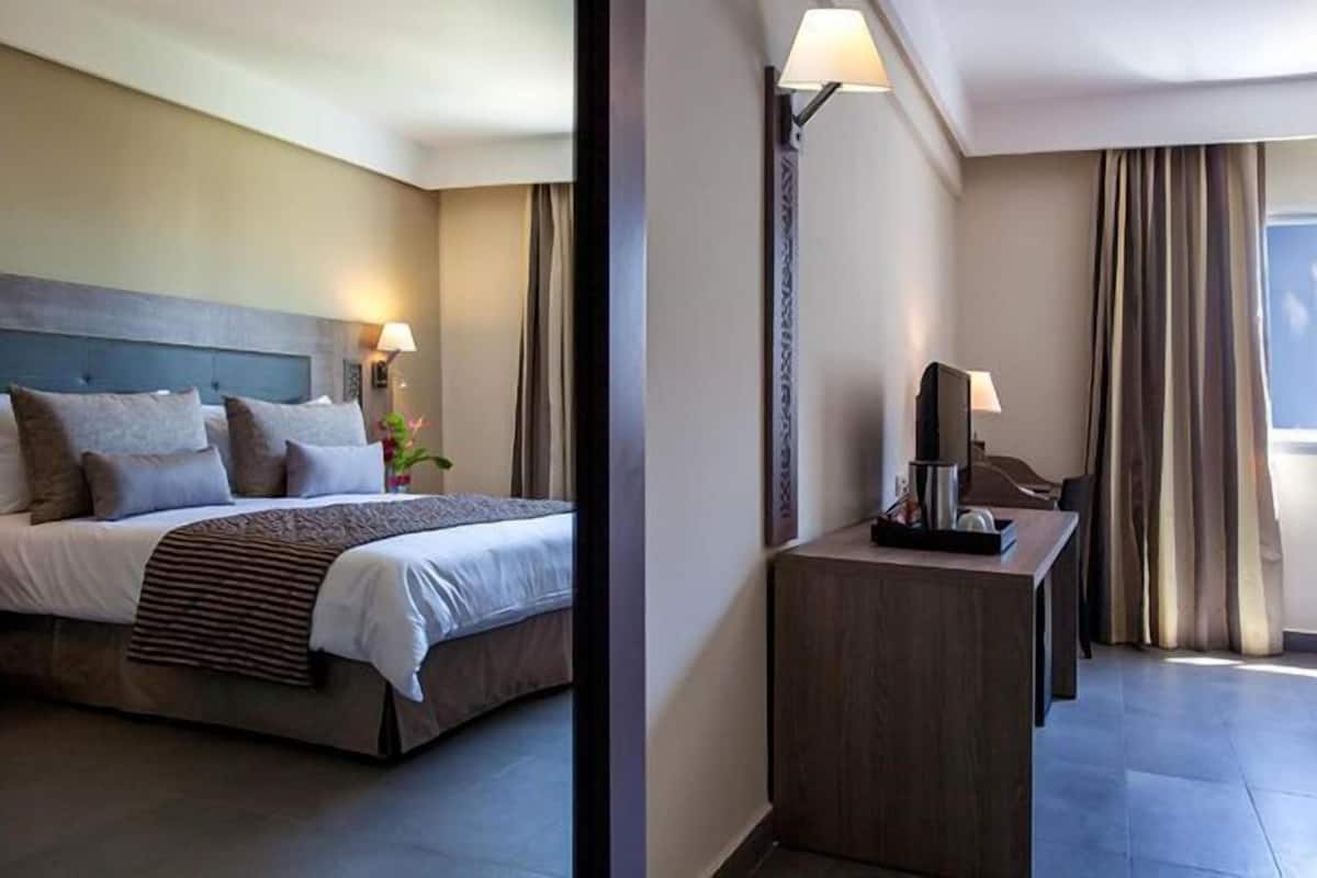 Quarto de hotel com uma cama grande, uma mesa de trabalho com luminária e uma janela com cortinas.