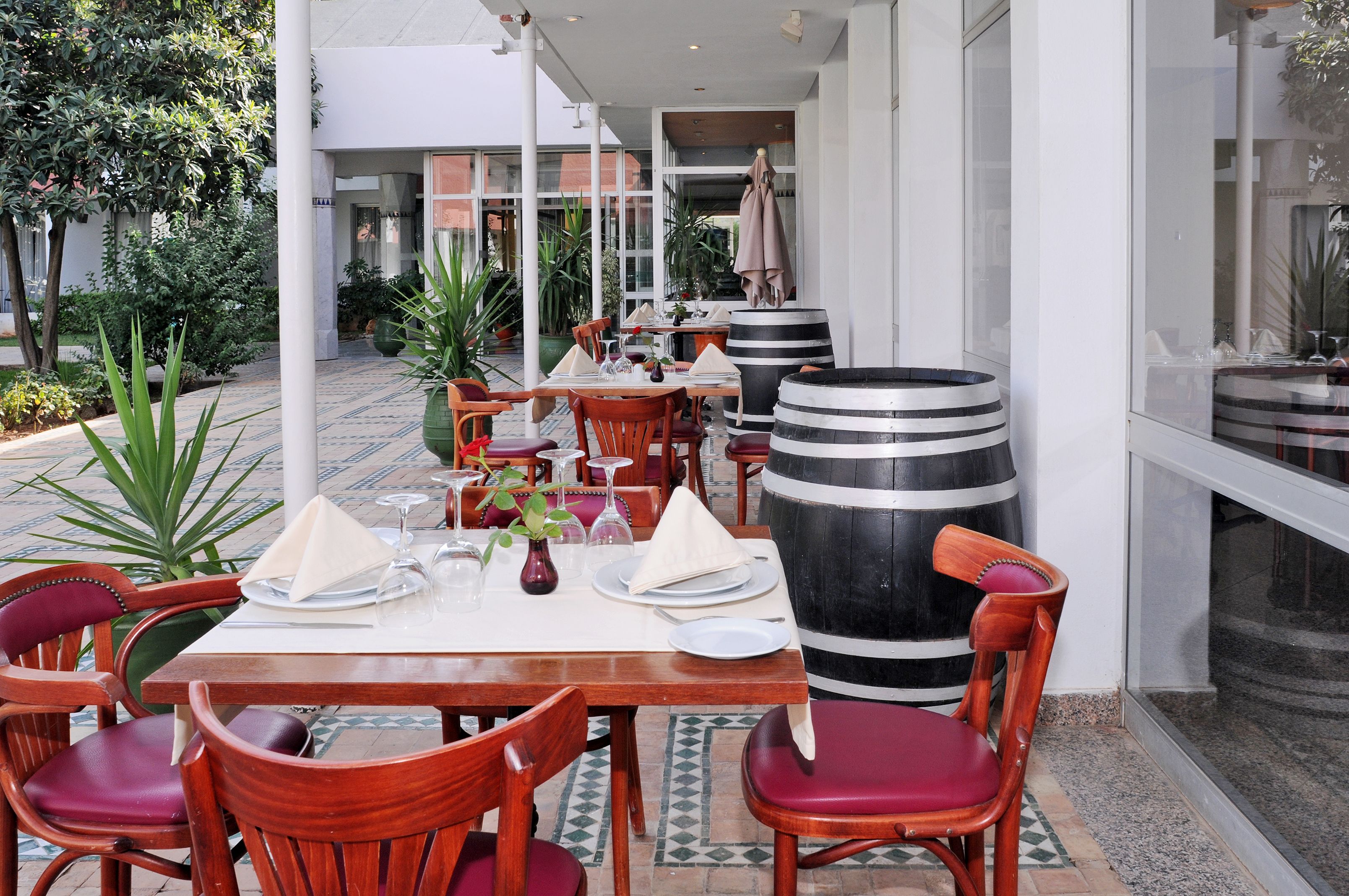 An outdoor dining area with wooden tables and chairs, a large black and white barrel, and potted plants.