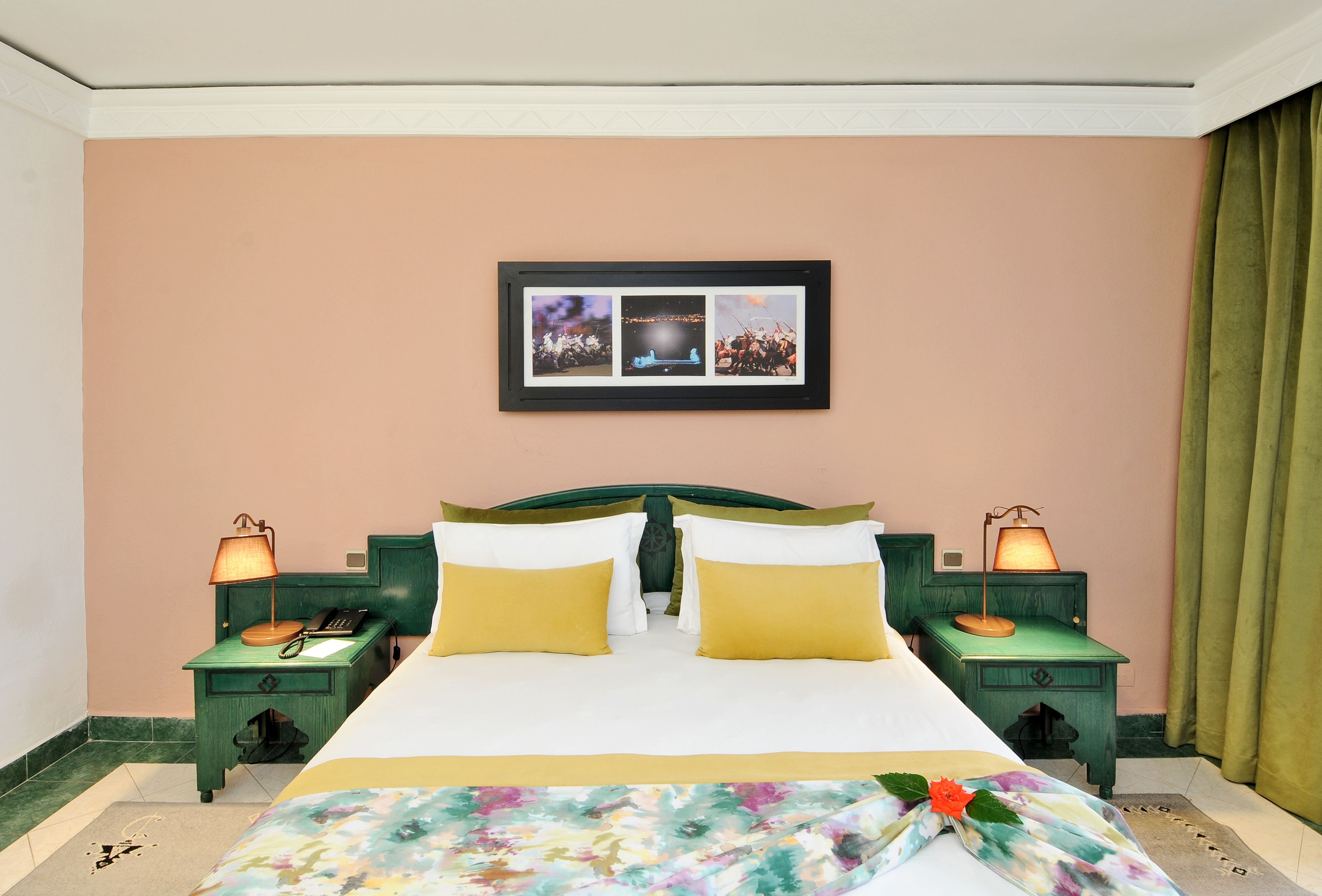 Habitación de hotel con una cama, dos mesitas de noche con lámparas, un cuadro enmarcado en la pared y una cortina verde.