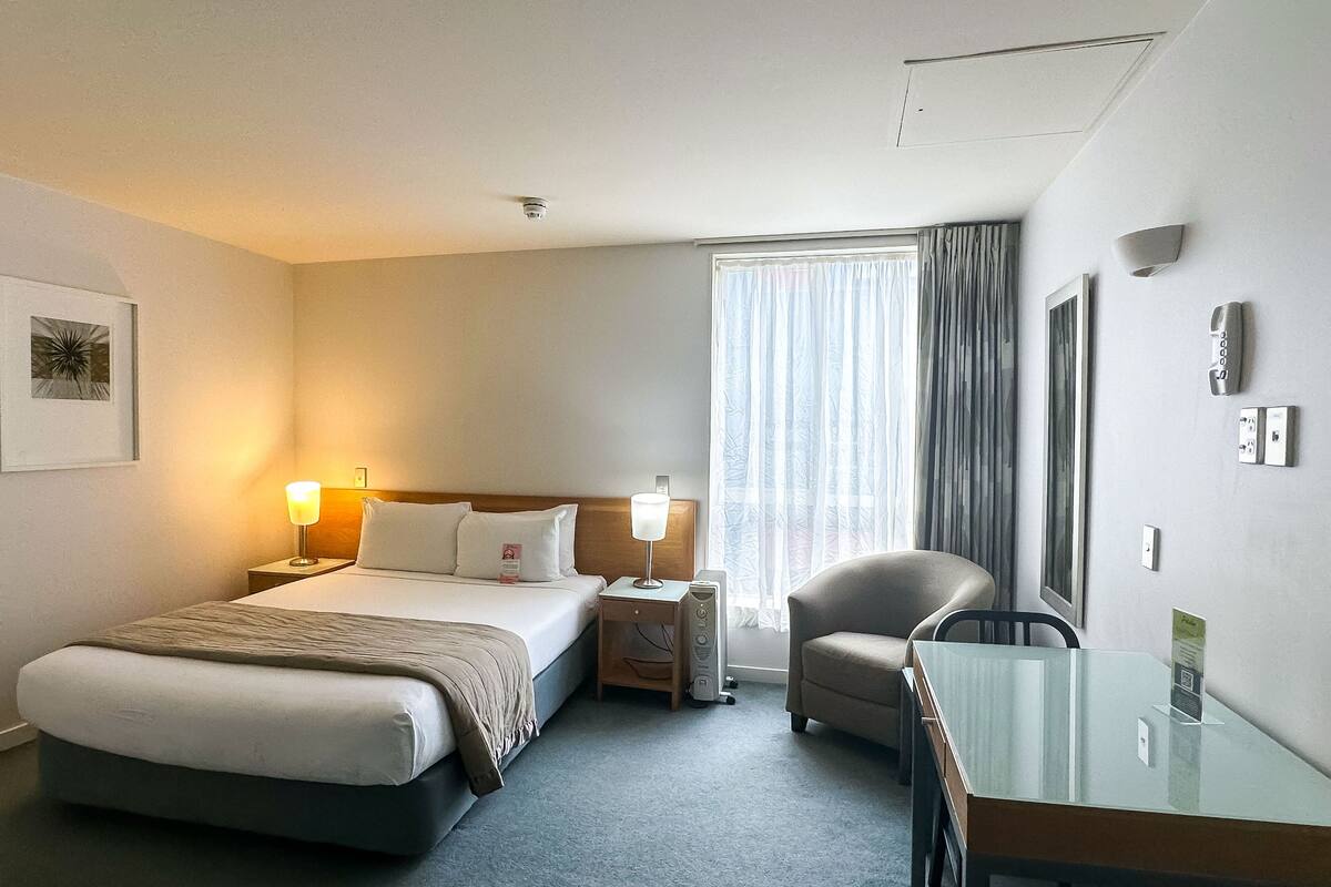 Een hotelkamer met een bed, bedlampen, een bureau, een stoel, een raam met gordijnen en een telefoon aan de muur.