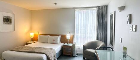 A hotel room with a bed, bedside lamps, a desk, a chair, a window with curtains, and a wall-mounted phone.
