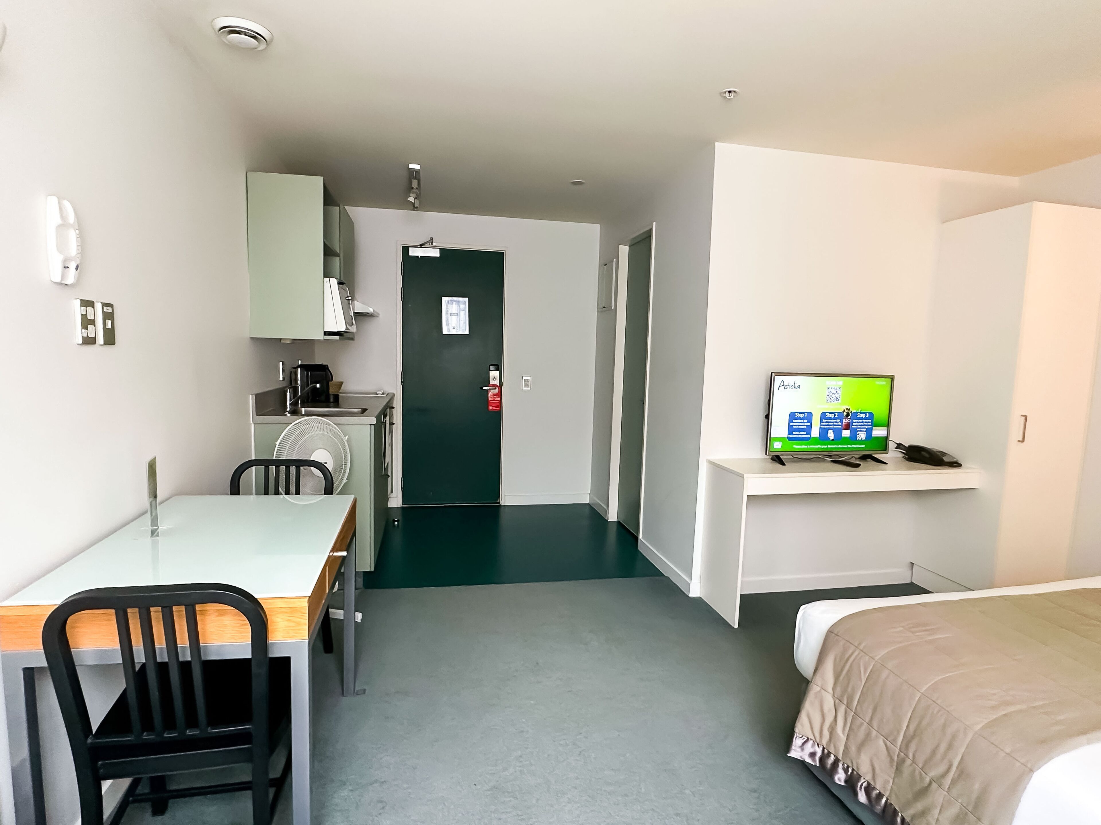 A hotel room with a bed, a desk with a chair, a television, a kitchenette, and a door.