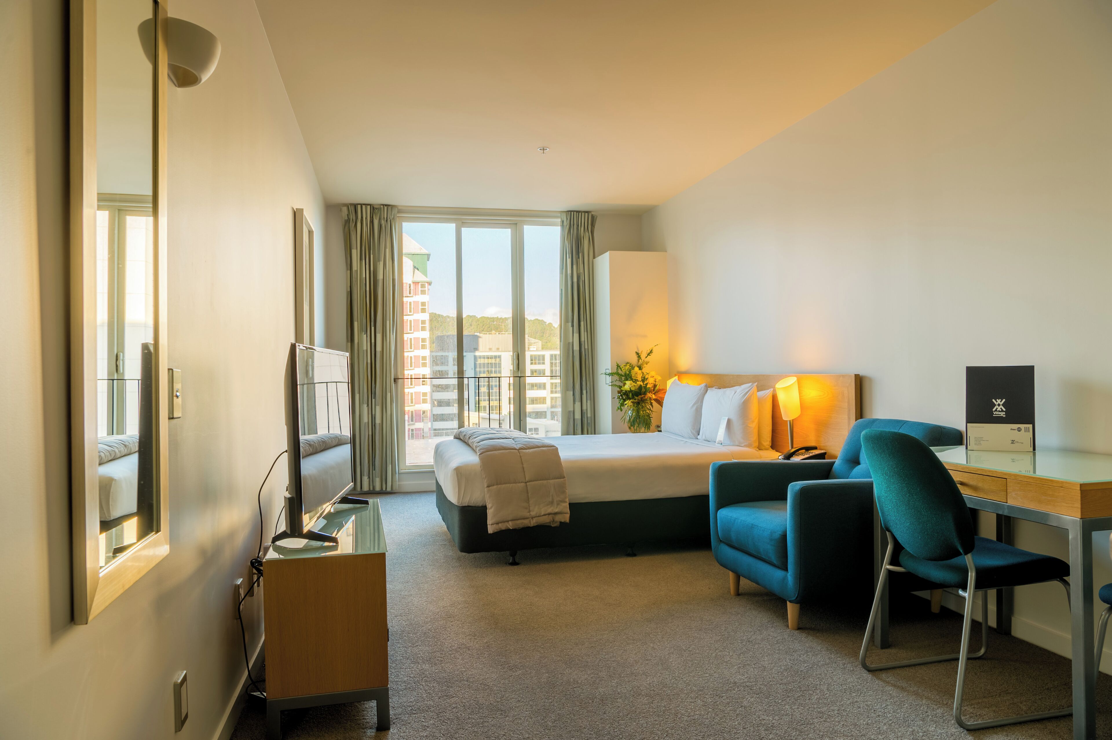 Quarto de hotel com cama, escrivaninha, cadeira, televisão e uma janela grande.
