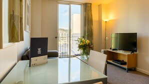 A hotel room with a glass table, a flat-screen TV on a wooden stand, a vase with flowers, and a view of buildings through a large window.