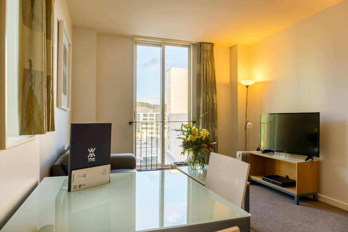 Een hotelkamer met een glazen tafel, een flatscreen televisie op een houten standaard, een bloemenvaas en een uitzicht op gebouwen door een groot raam.