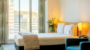 A hotel room with a large bed, a bedside table with a lamp, a chair, and a view of buildings through the window.