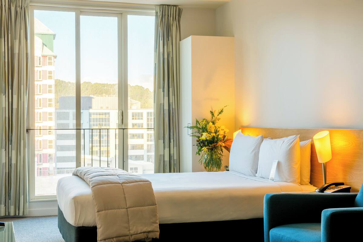 Een hotelkamer met een groot bed, een nachtkastje met een lamp, een stoel en uitzicht op gebouwen door het raam.