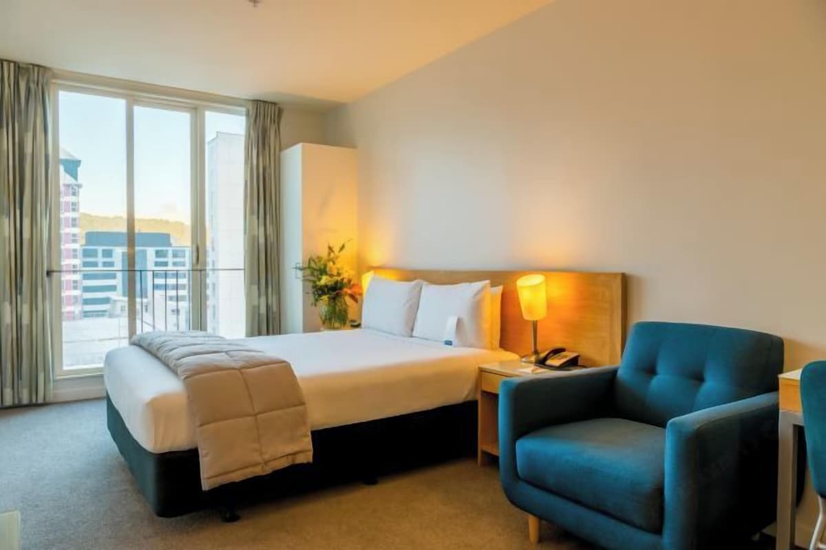 Habitación de hotel con una cama grande, un escritorio, una silla y vistas a edificios a través de la ventana.
