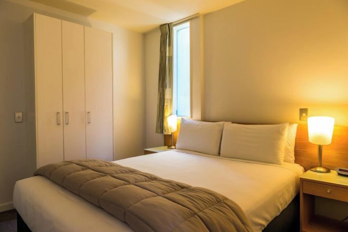 Habitación de hotel con cama, mesita de noche con lámpara, armario y ventana con cortinas.
