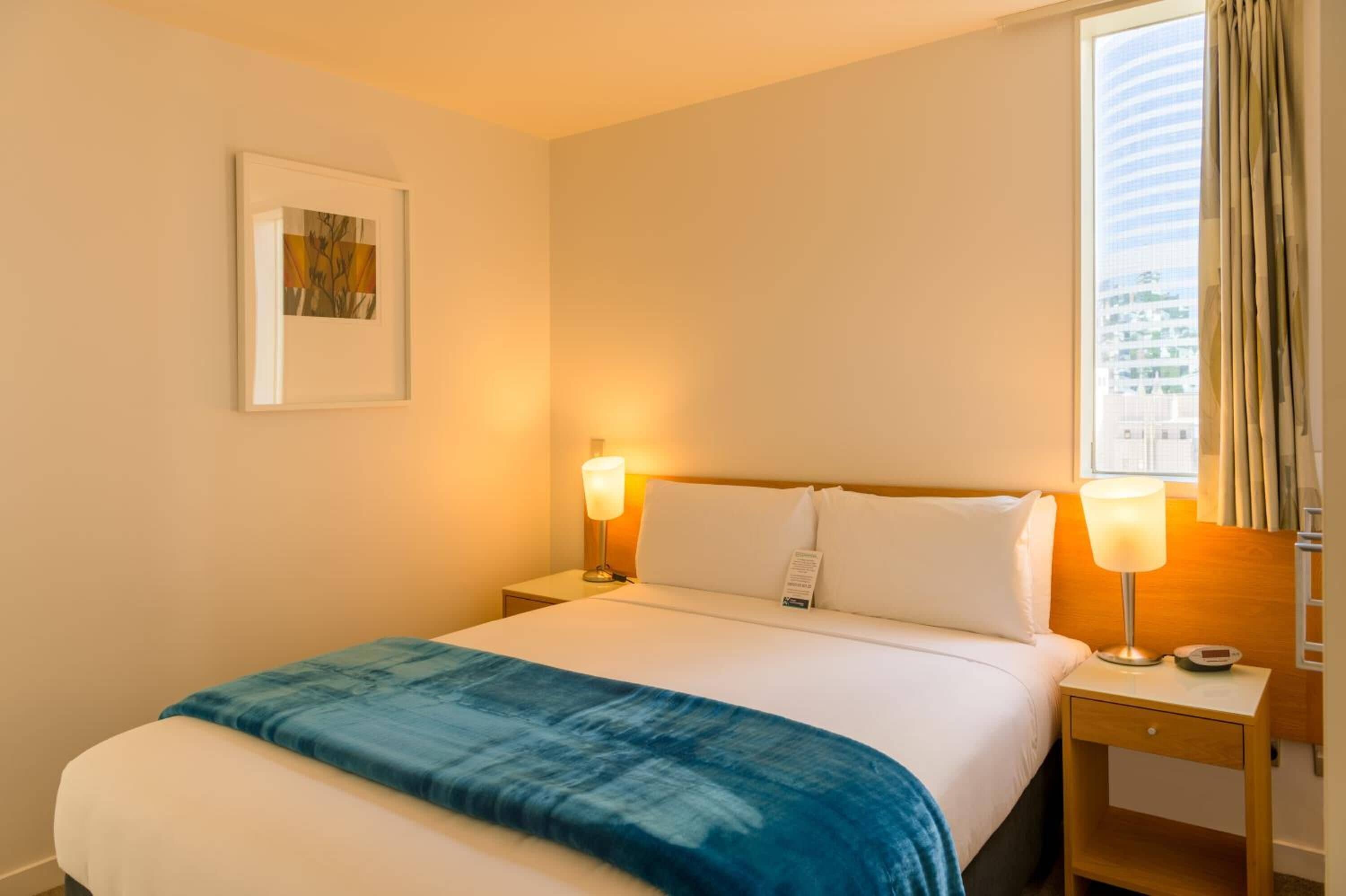 Hotellihuone, jossa on sänky, kaksi yöpöytälamppua, yöpöytä, seinällä oleva kehystetty kuva ja ikkuna, jossa on verhot.