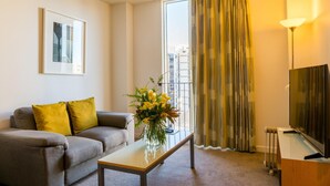 A modern living room with a sofa, a coffee table with a vase of flowers, a flat-screen TV, and a large window with curtains.