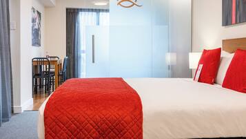 A hotel room with a large bed, a glass partition wall, a wooden desk with chairs, and a red and white color scheme.