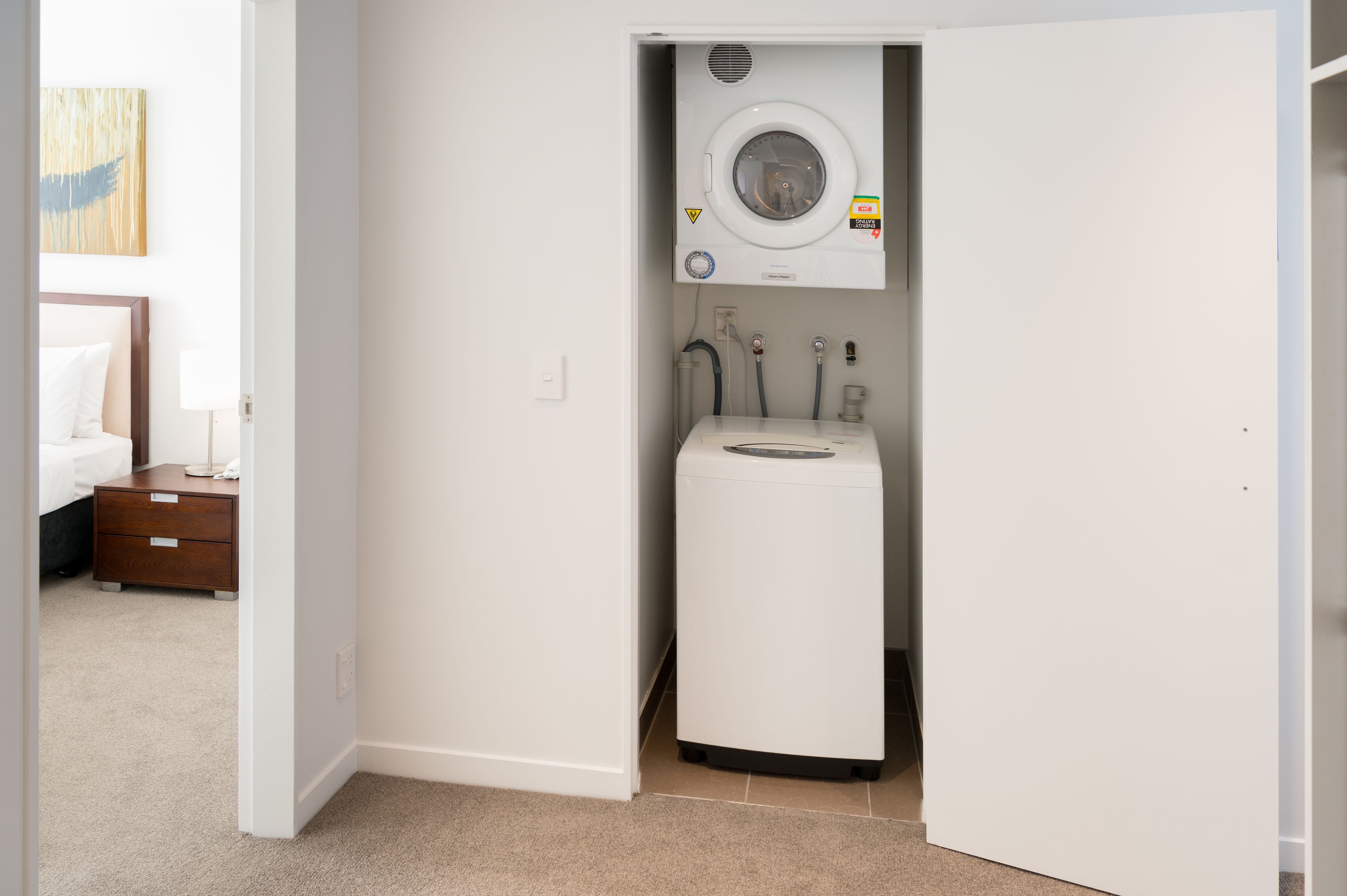 A compact laundry room with a front-loading washer and dryer, a small cabinet, and a bed in the adjacent room.