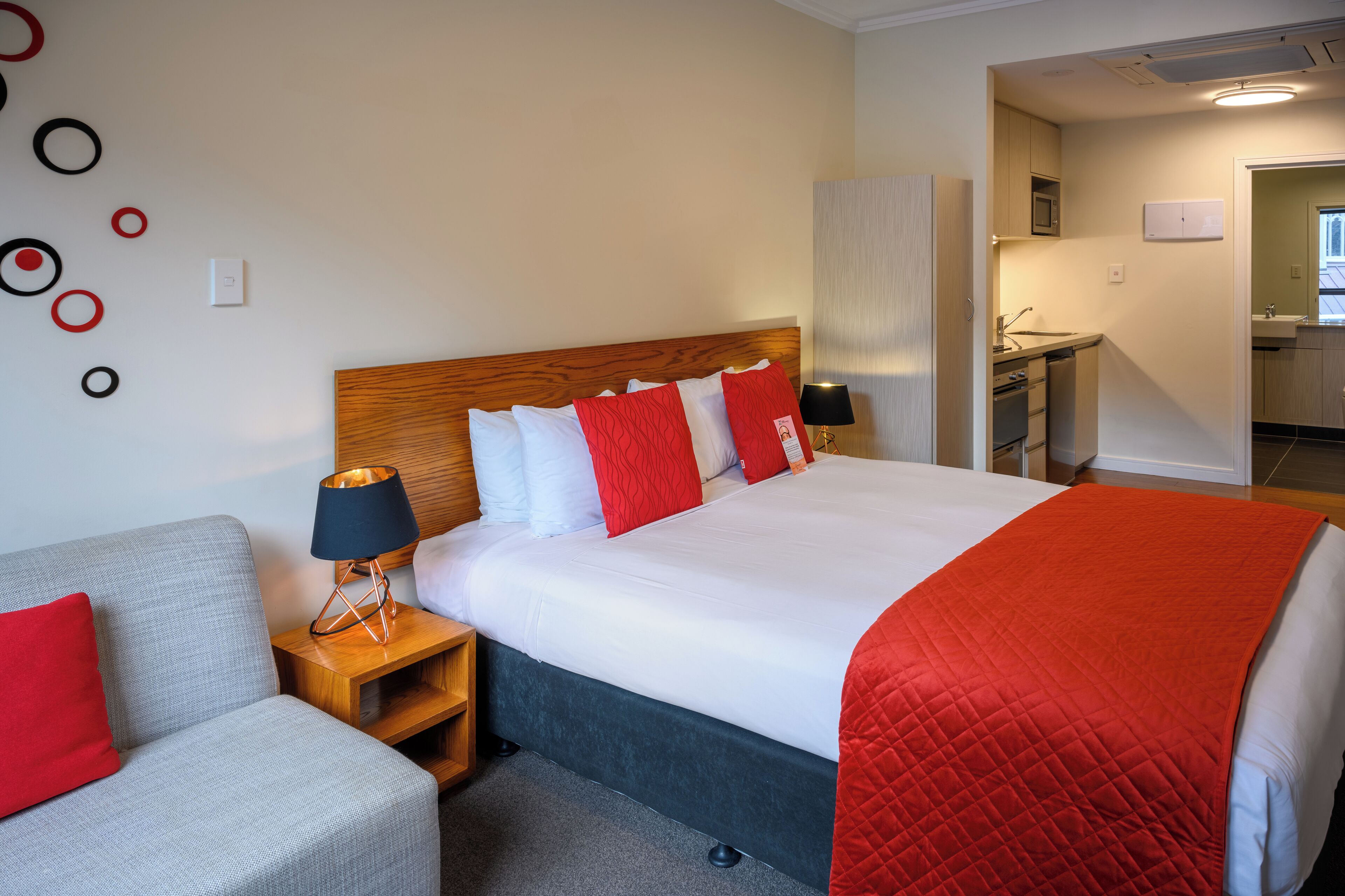 A hotel room with a bed, a grey sofa, a wooden nightstand with a lamp, and a red quilted blanket.