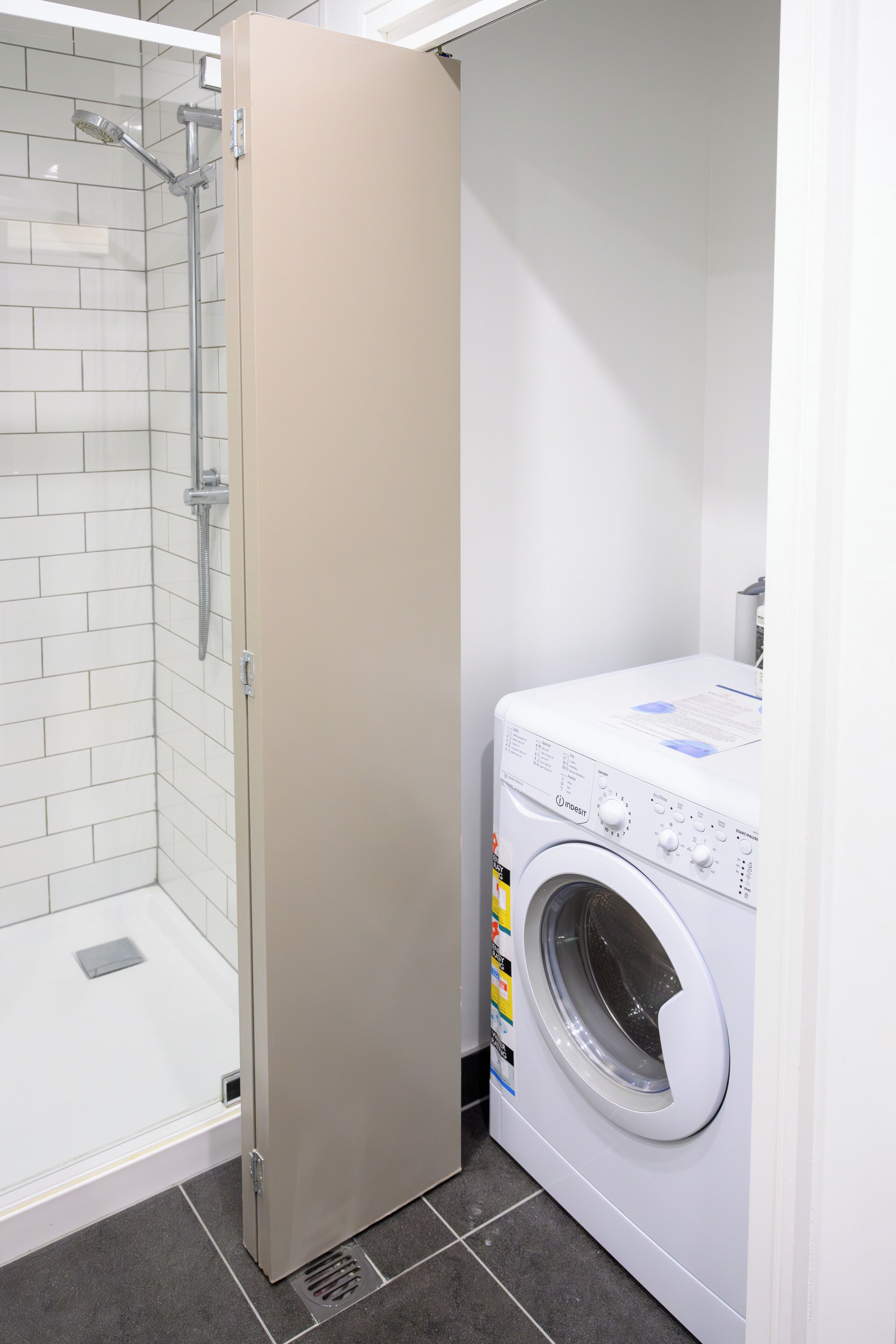 A compact bathroom with a glass shower enclosure, a washing machine, and a beige door.