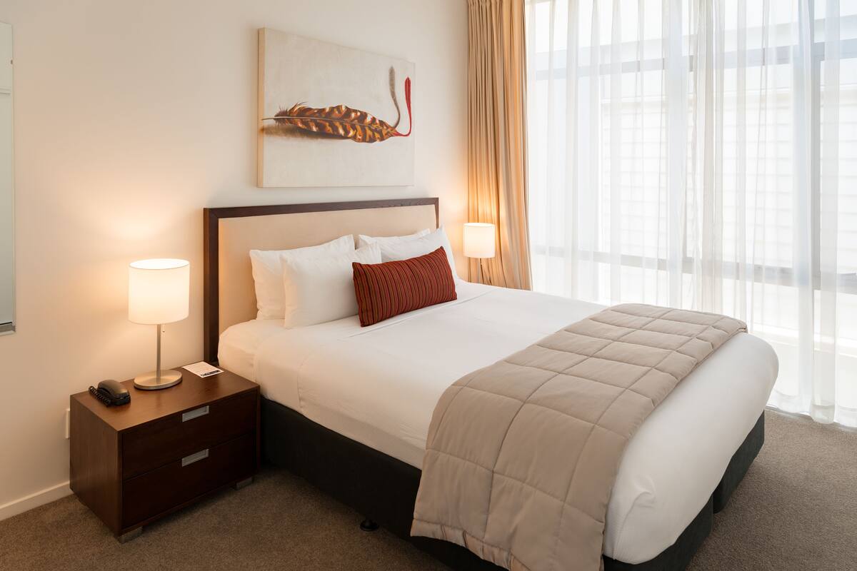 Une chambre d’hôtel avec un grand lit, deux lampes de chevet, une table de chevet, une œuvre d’art encadrée au-dessus du lit et des rideaux légers à la fenêtre.