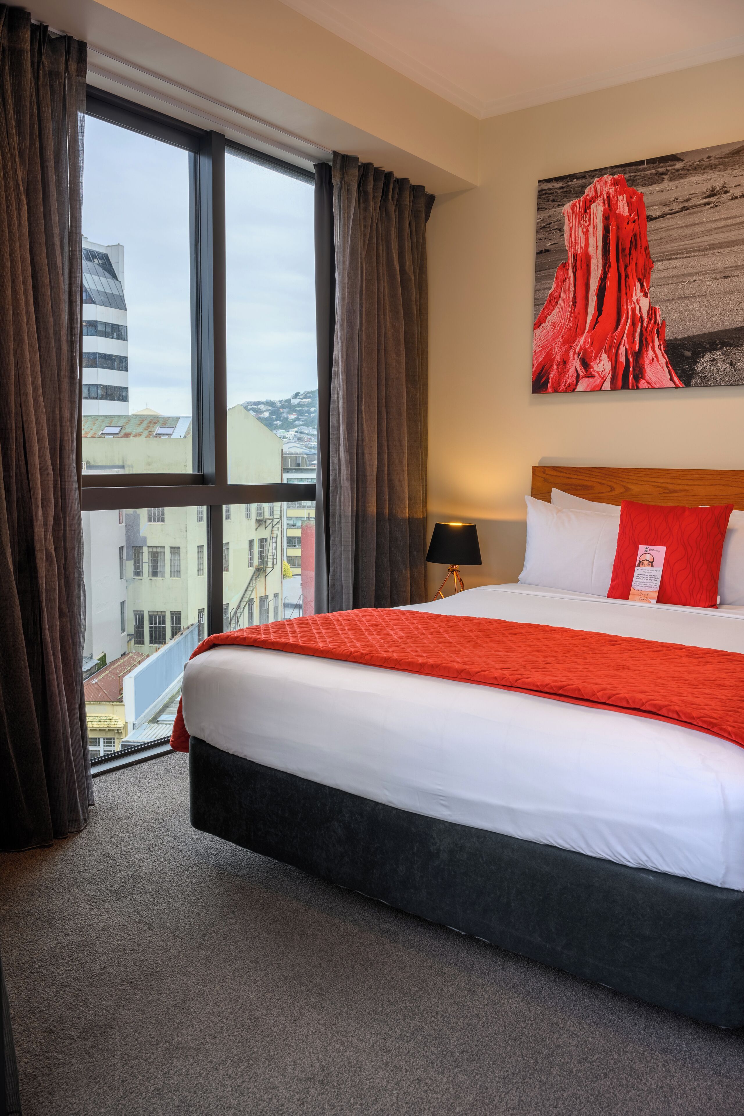 Una camera d'hotel con un letto grande, vista sulla città dalla finestra, un quadro appeso al muro e una lampada sul comodino.