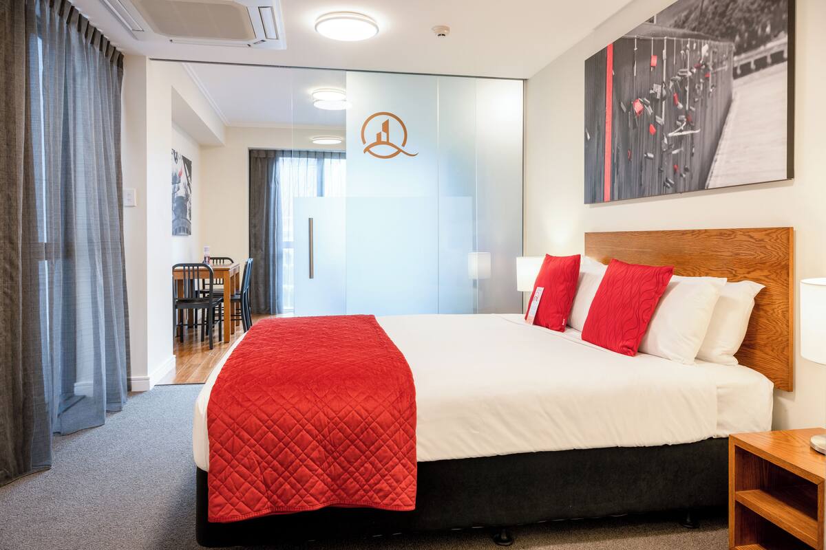 Une chambre d’hôtel avec un grand lit, une tête de lit en bois, une couvre-lit rouge matelassée et un oreiller rouge. Il y a un tableau au mur et un bureau avec une lampe.
