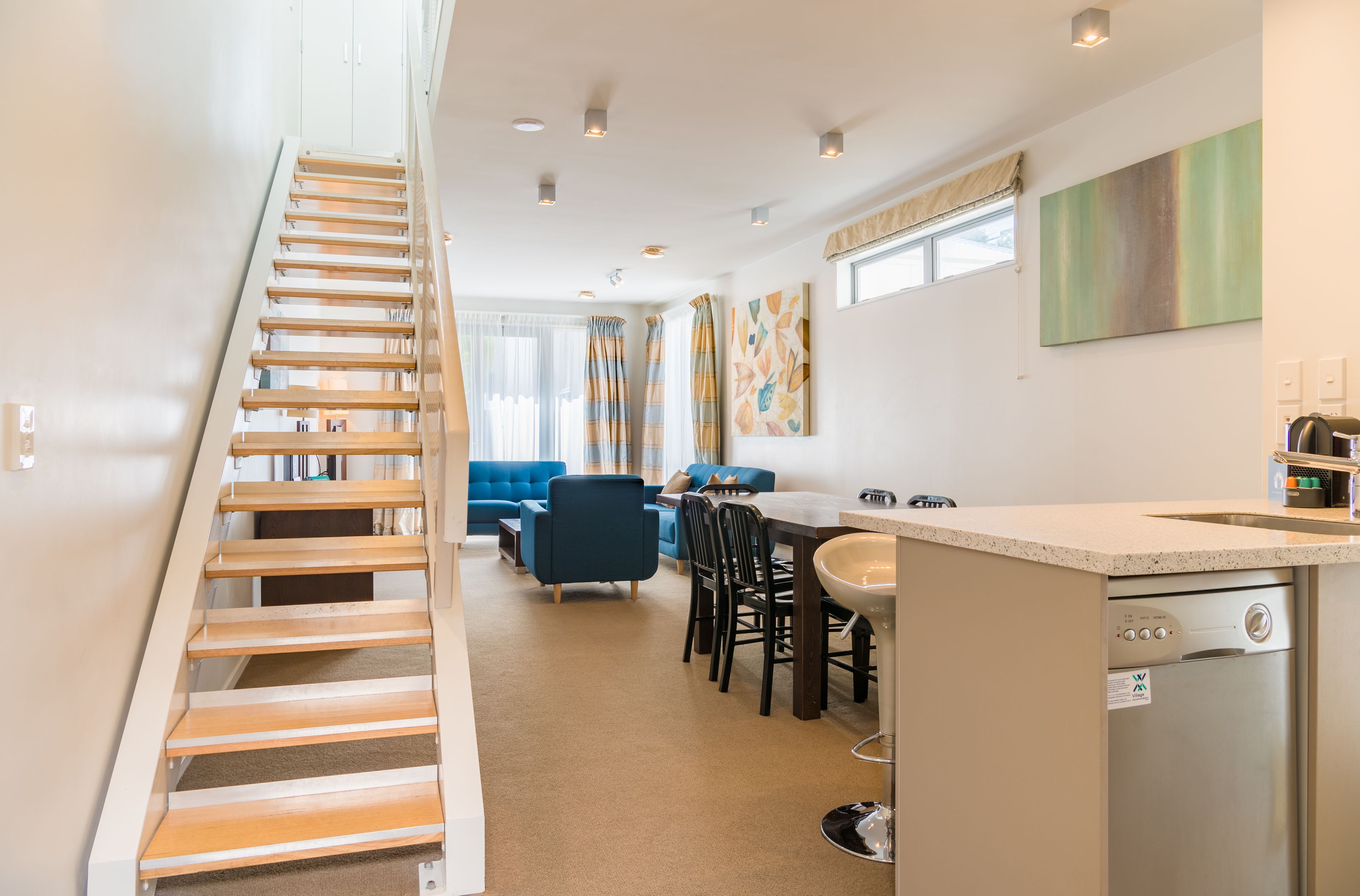 A modern kitchen with a dishwasher, a dining area with chairs and a table, and a staircase leading to another level.