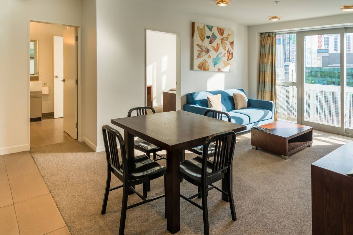 Un salon moderne comprenant une table à manger, des chaises, un canapé et une grande fenêtre donnant sur des immeubles.