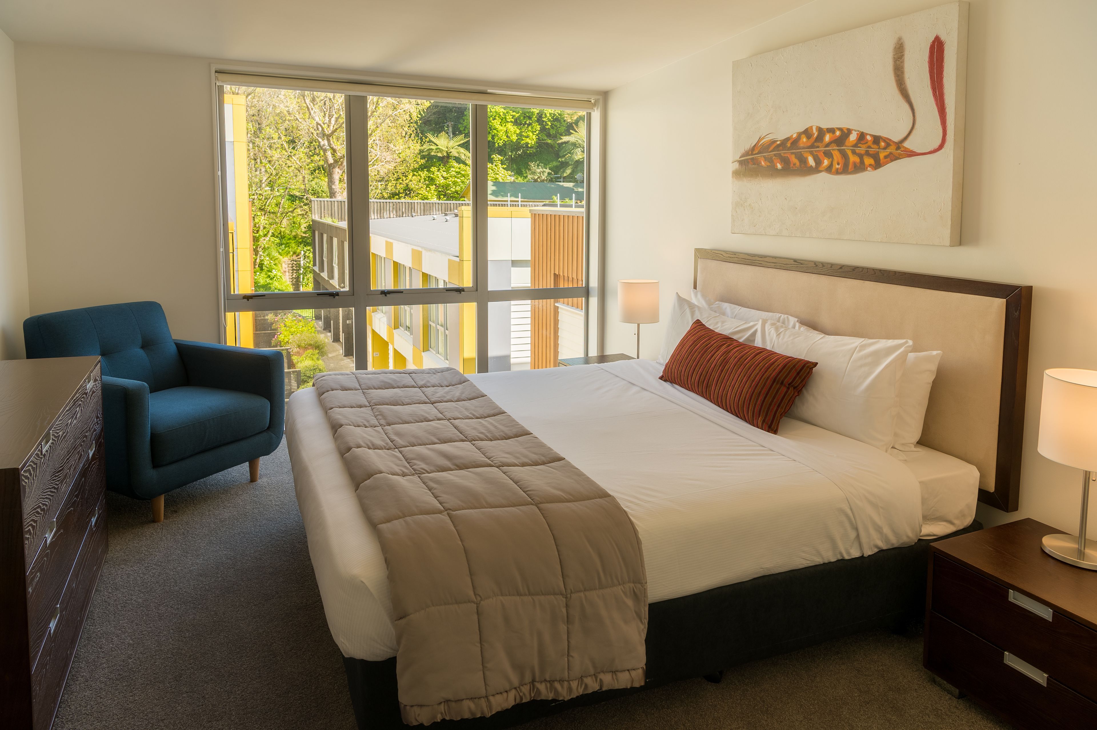 A hotel room with a large bed, a blue armchair, a wooden nightstand, and a window with a view of greenery and buildings.